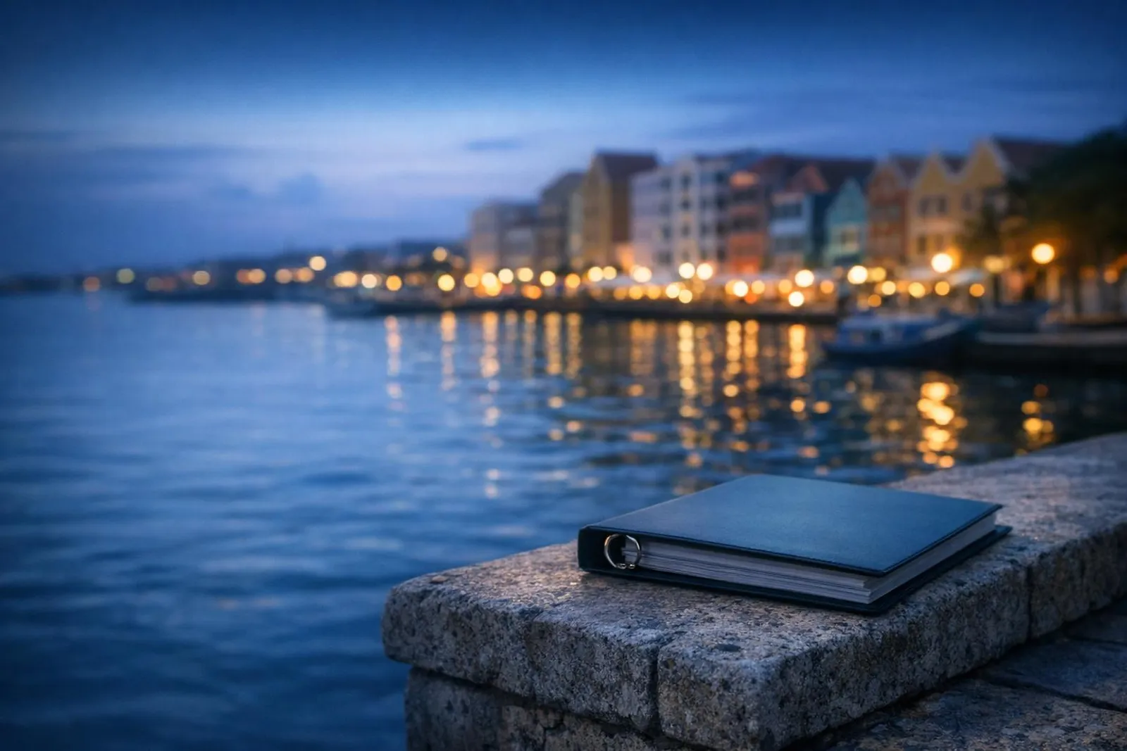 Quiet Caribbean harbour at dusk, colourful waterfront facades softly blurred, closed ring binder resting on a stone ledge in 